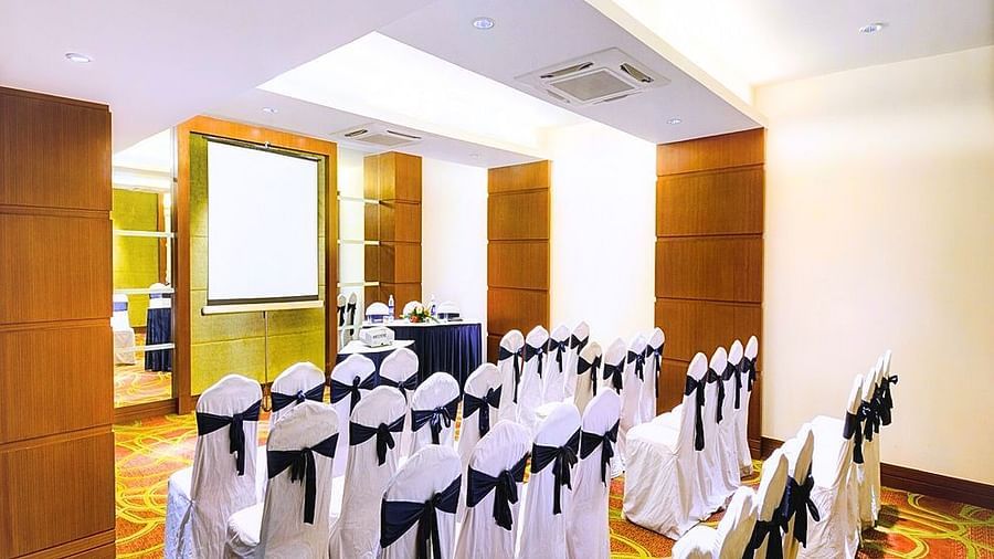 A conference room at Pride Plaza Ahmedabad, featuring rows of chairs with black sashes, a projector screen, and natural light streaming in.